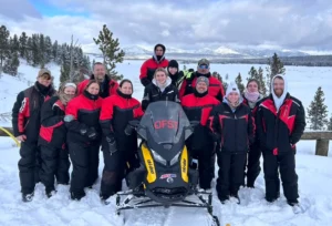 Group of snowmobilers in red and black winter gear posing with yellow snowmobile in snowy mountain landscape with forests ...
