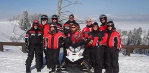 Group of snowmobilers in red and black winter gear posing with white snowmobile on snowy mountain landscape
