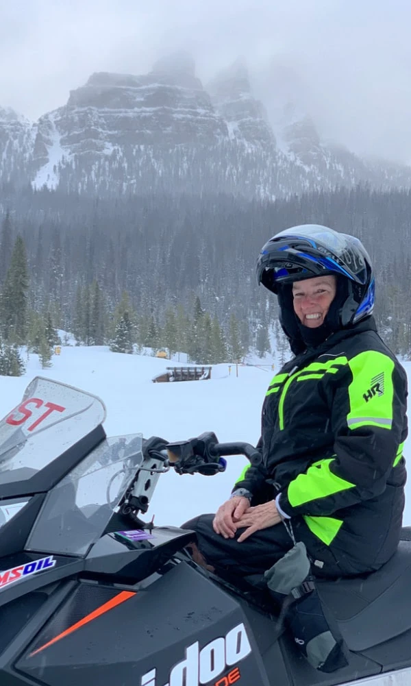 Person in bright green jacket and helmet smiling on snowmobile in snowy mountain landscape with forest and rocky peaks in ...
