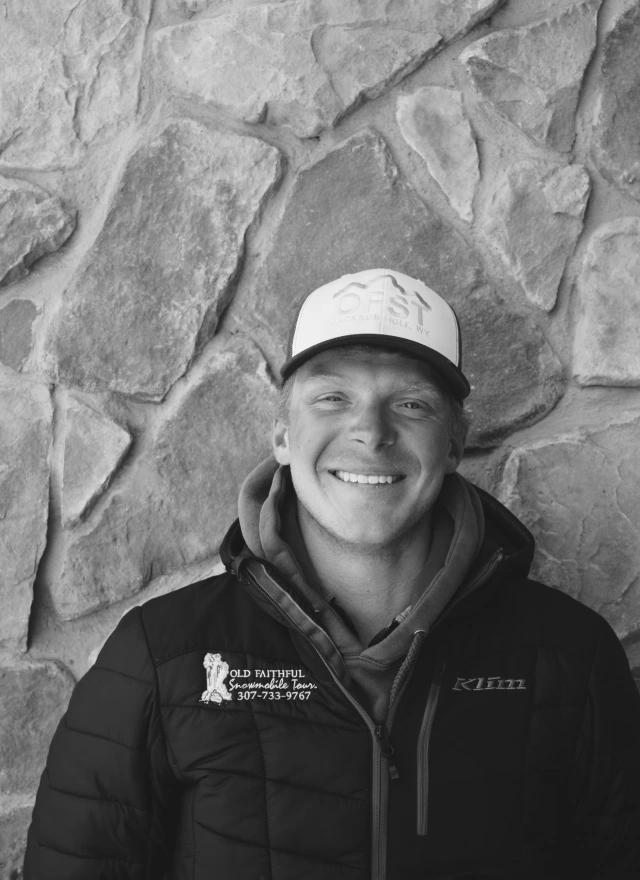 Professional guide Owen Lynch smiling in black winter jacket and cap against stone wall background