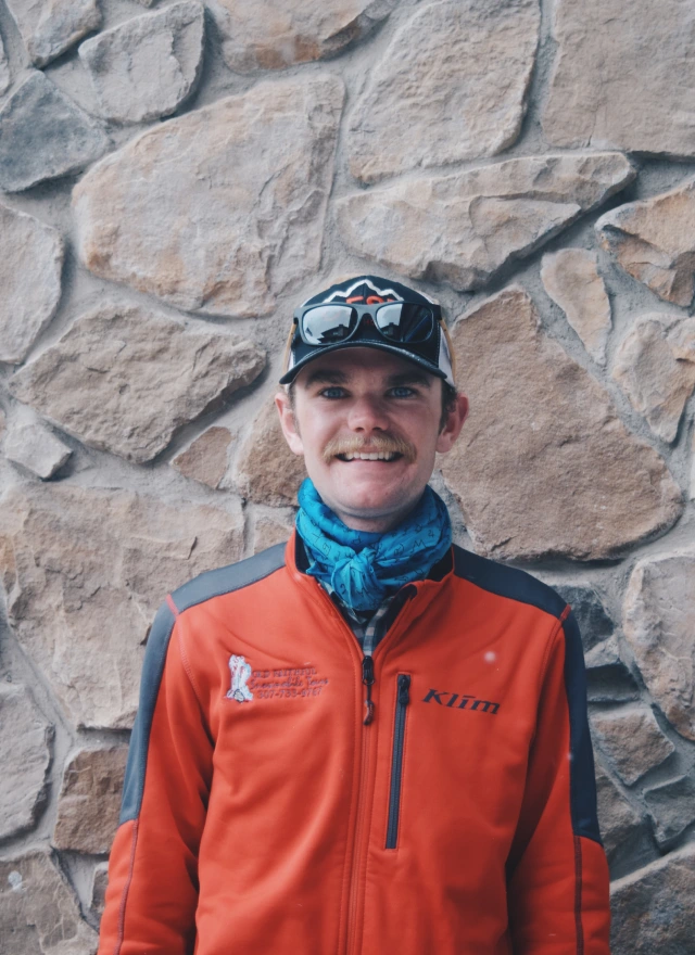 Guide Peyton Moore wearing orange outdoor jacket and cap, smiling in front of stone wall