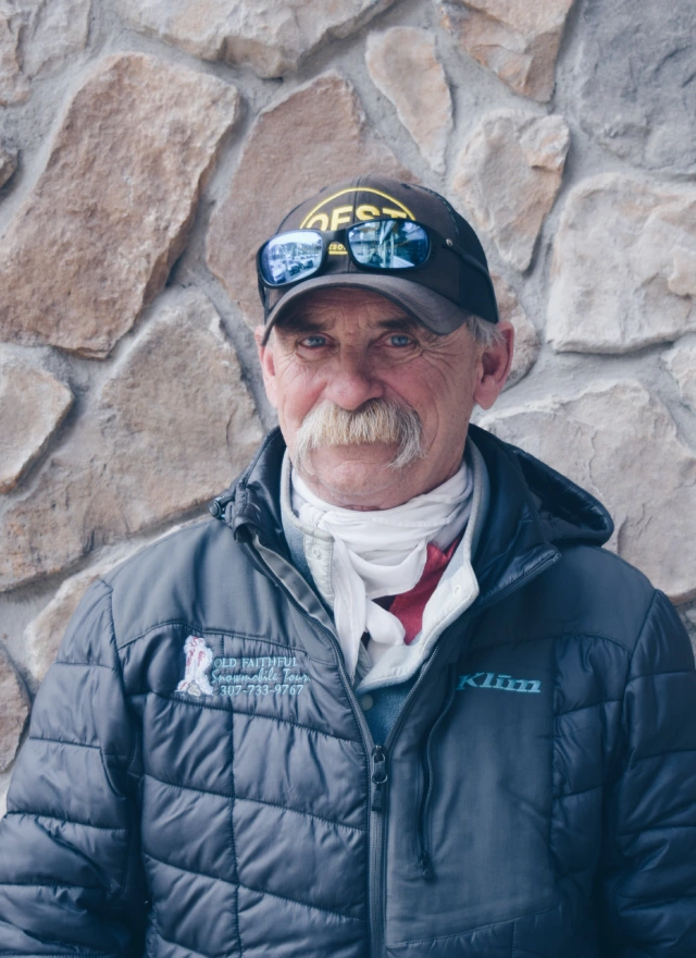 Older man with mustache wearing puffy jacket and baseball cap, standing against stone wall background