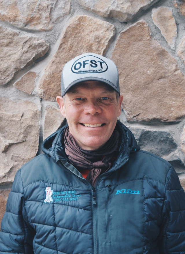 Man in puffy jacket and baseball cap smiling at camera against stone wall background