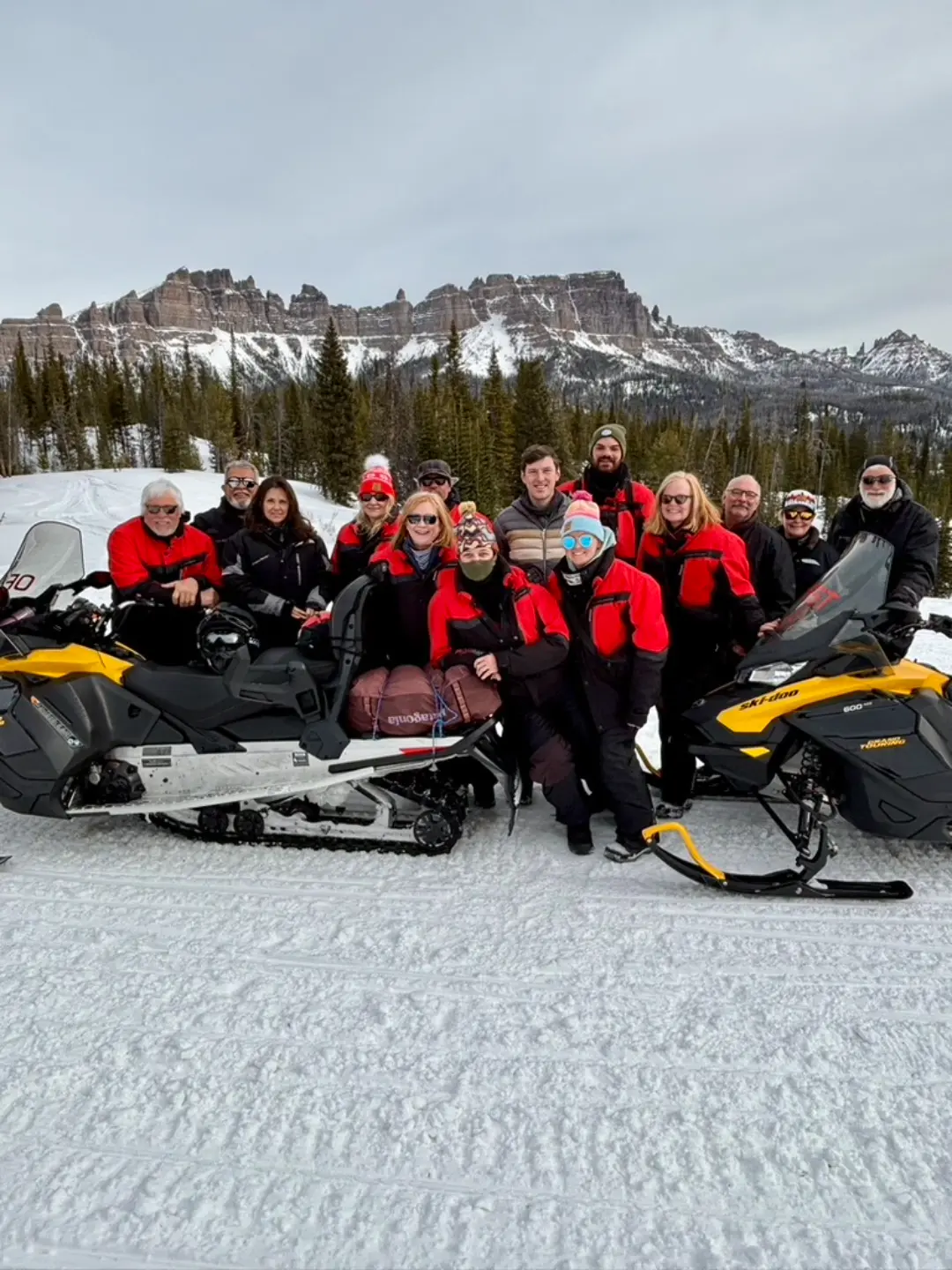 Group of snowmobilers posing with their machines in front of snow-covered Rocky Mountains during Continental Divide tour
