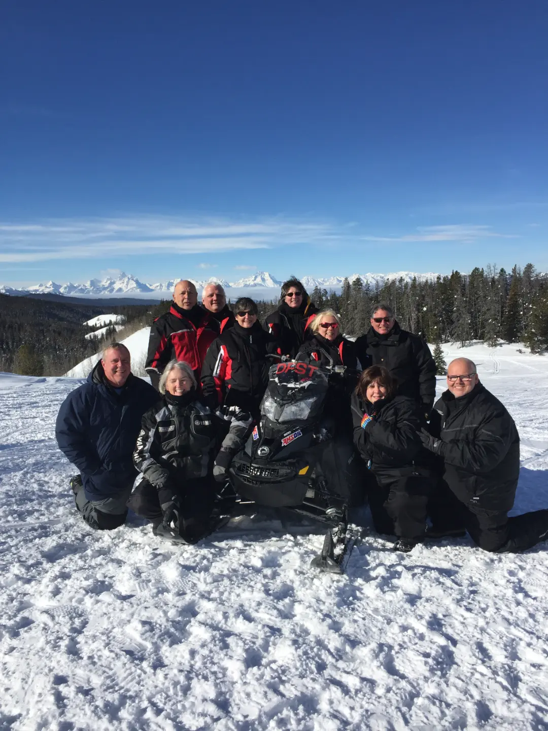 Group of snowmobilers posing with black snowmobile in snowy mountain landscape with evergreen forests and snow-capped peak...