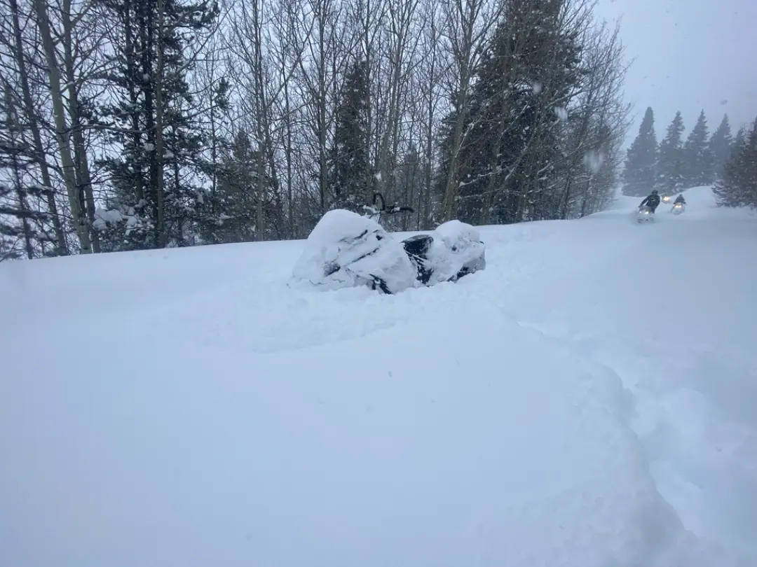 Snow-covered rocks on snowy slope with snowmobilers in background among evergreen trees during Continental Divide tour