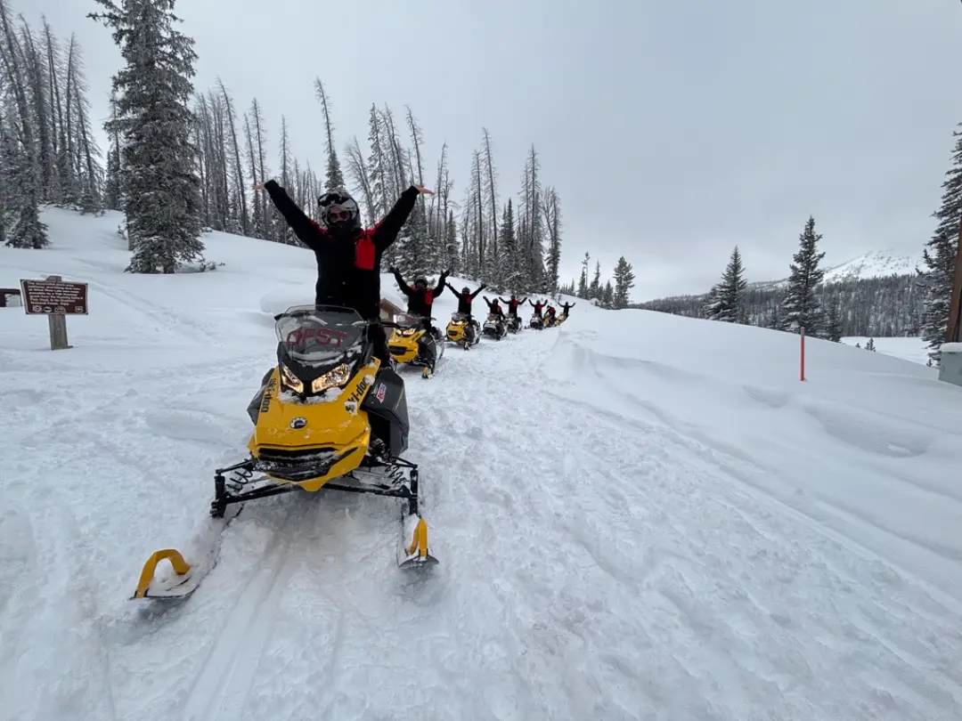 Group of snowmobilers with raised arms celebrating on snowy trail through mountain forest during Continental Divide tour