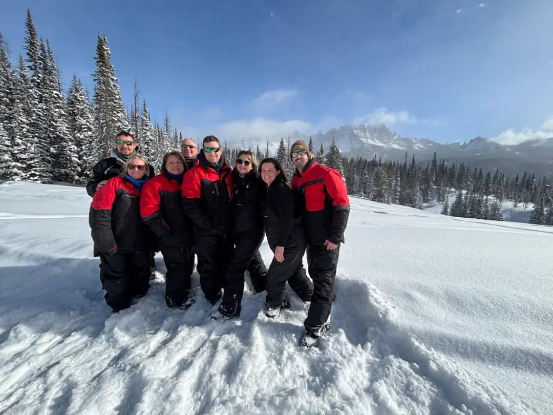 Group of eight snowmobilers in red and black winter gear posing in deep snow with snow-covered evergreen trees and mountai...