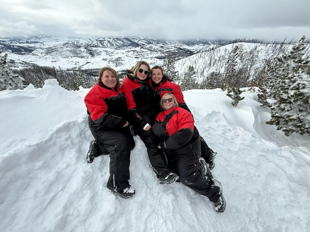 Four people in red and black winter gear sitting in snow with snowshoes, mountains and forest visible in background on Con...