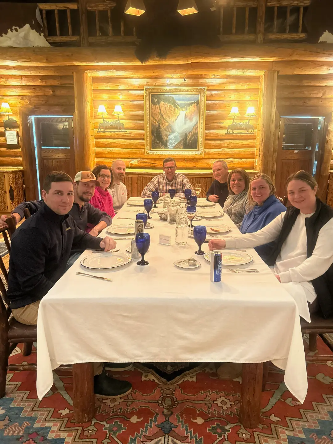 Family group dining together at rustic log cabin restaurant with white tablecloth, blue glasses, and landscape painting on...