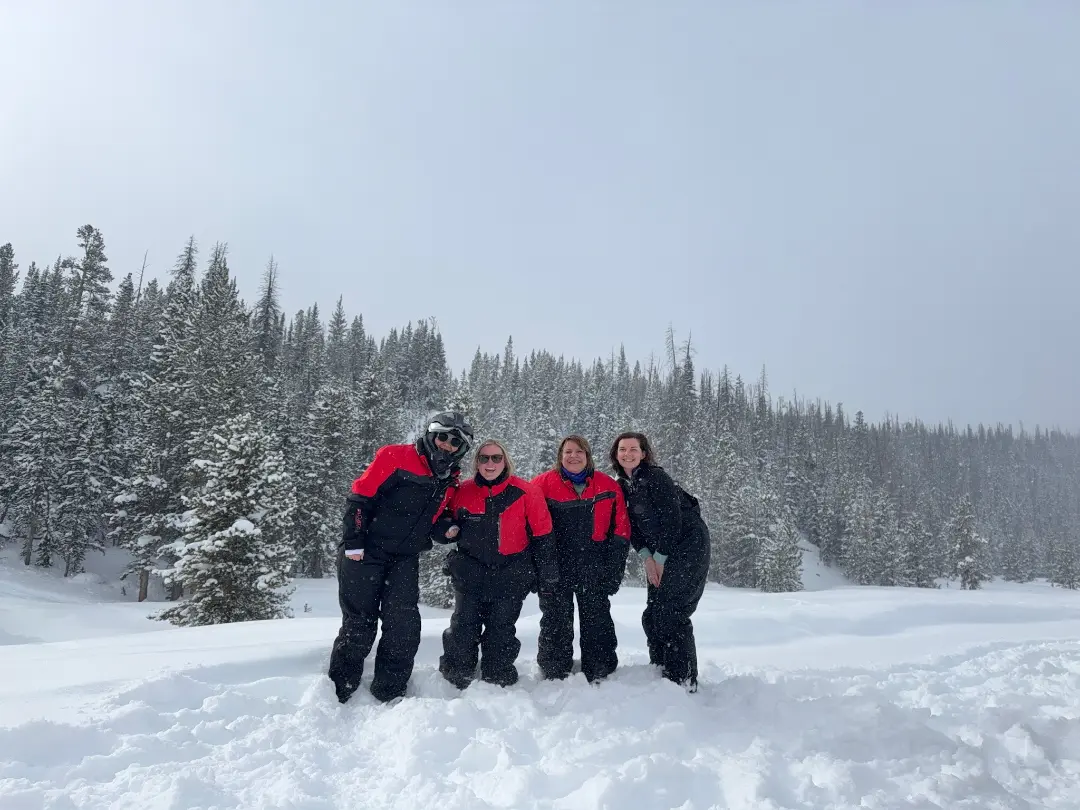 Four people in winter gear pose together in deep snow during a snowmobile tour, with snow-covered evergreen trees and moun...