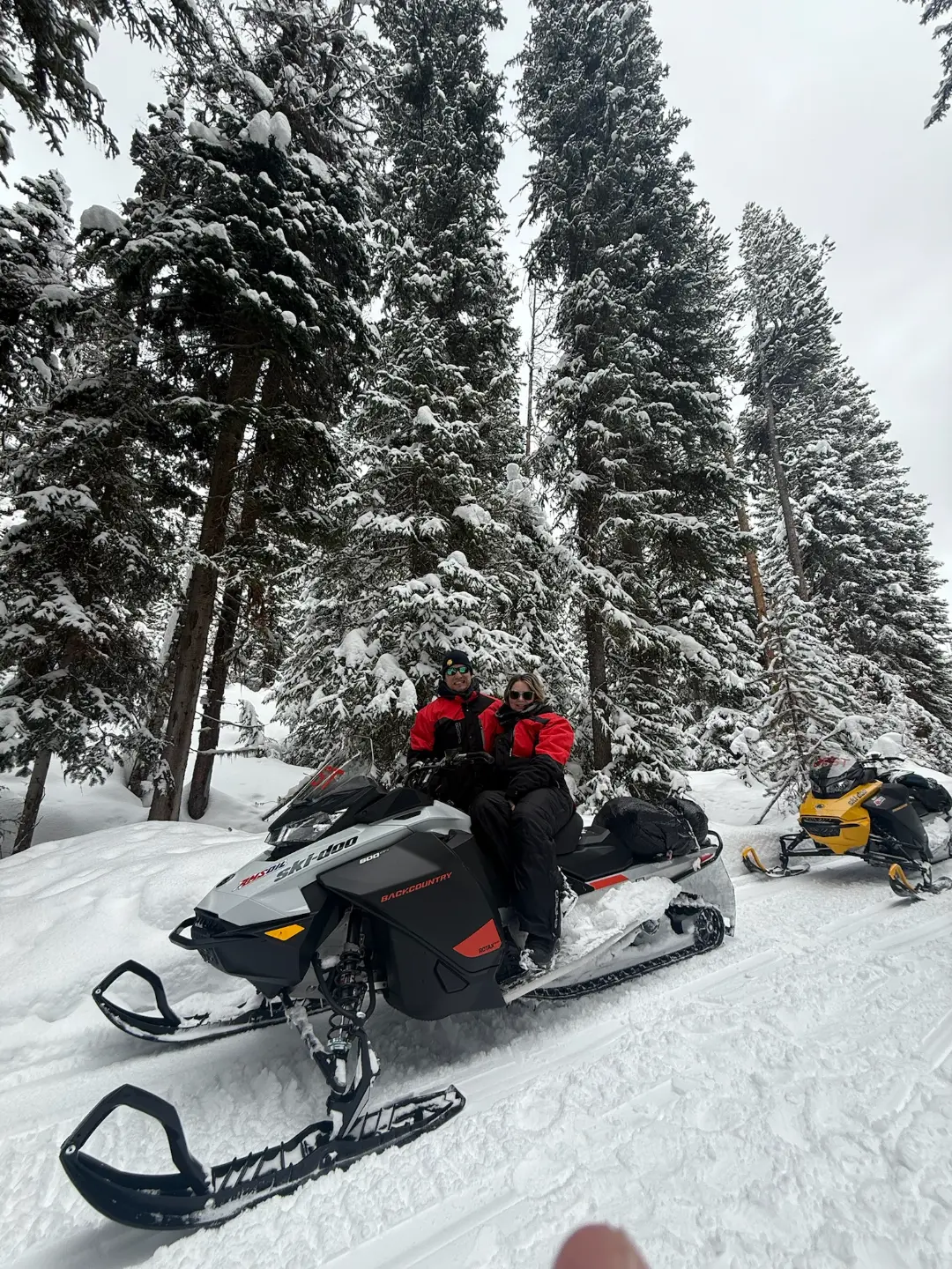 Two snowmobilers in red jackets sitting on a snowmobile surrounded by snow-covered evergreen trees during a Continental Di...