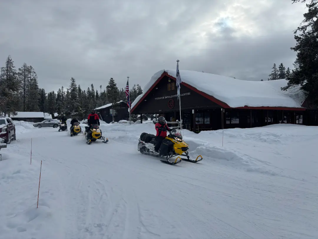 Snowmobilers gathered at Turquoise Meadow Ranch lodge during Continental Divide tour, with yellow snowmobiles parked in de...