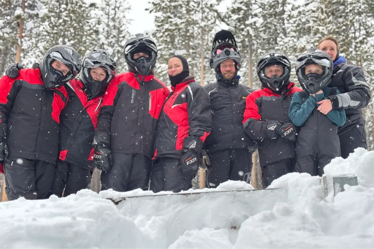 Group of people in winter gear and helmets posing together in snowy forest during snowmobile tour