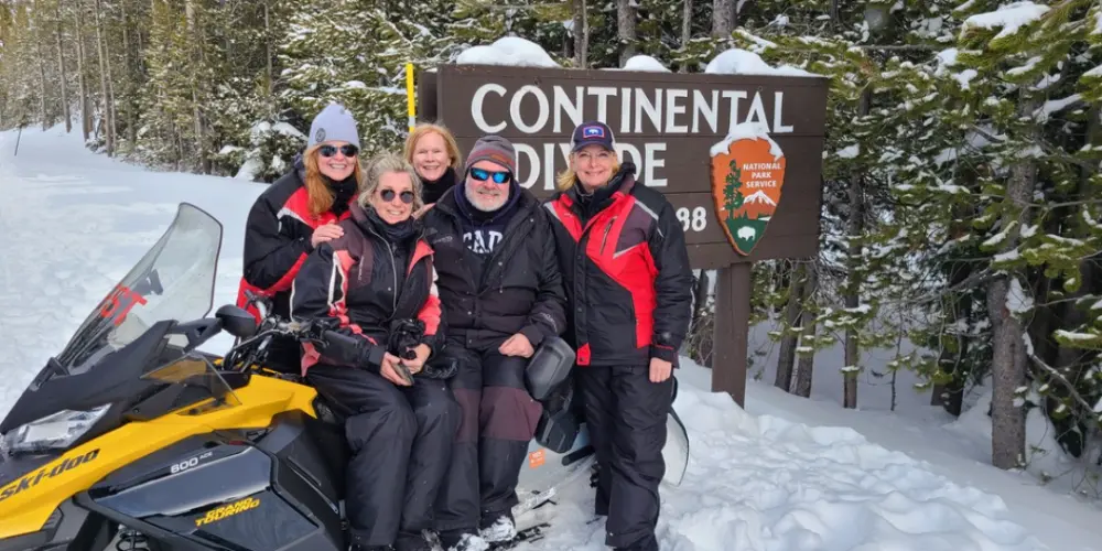 Group of five people in winter gear posing with snowmobile at Continental Divide sign in snowy forest during Yellowstone tour
