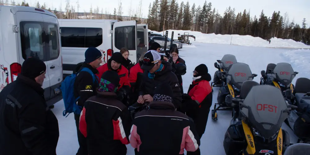 Group of people in winter gear gathered around someone on snowy ground near snowmobiles and white van in forest setting