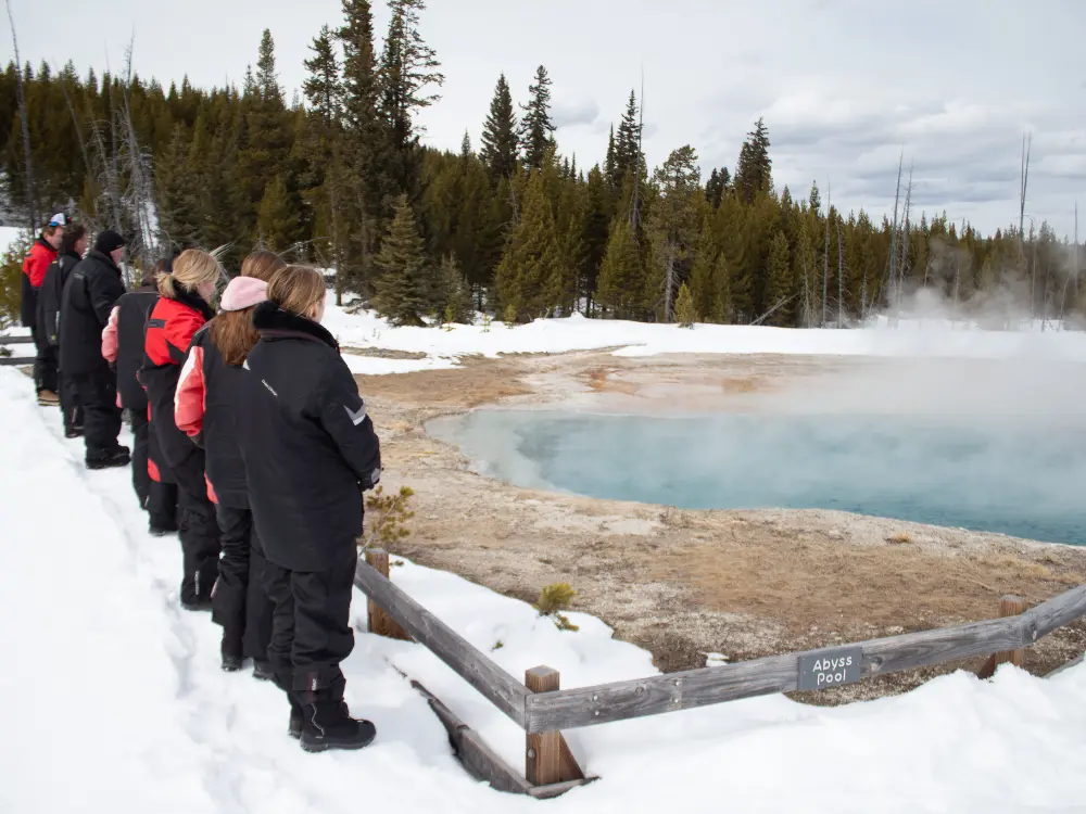 Tourists in winter gear viewing steaming Abyss Pool hot spring in snowy Yellowstone National Park during snowmobile tour