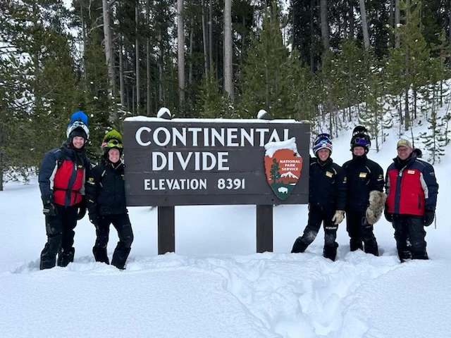 Group of snowmobile tour guides posing by Continental Divide elevation 8391 sign in snowy forest setting
