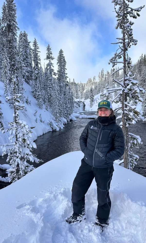 Person in winter gear standing in deep snow beside a flowing river surrounded by snow-covered evergreen trees in Yellowstone